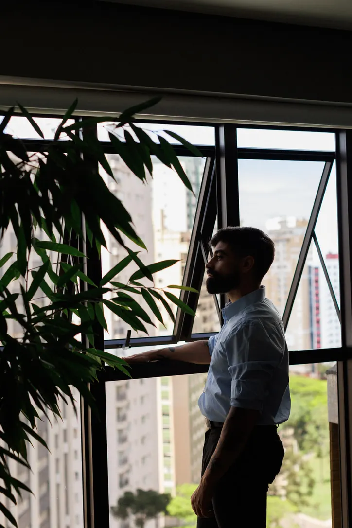 Júlio à janela do consultório, com plantas e a vista da cidade